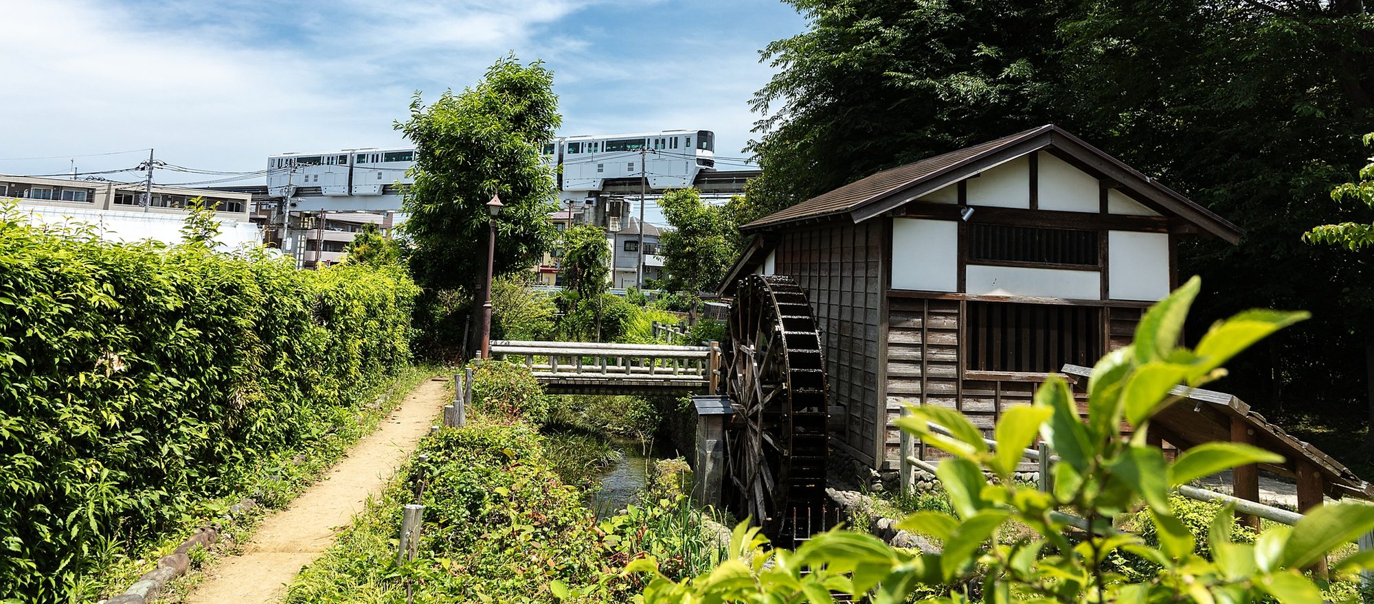 日野市の風景。用水路沿いの小径と水車小屋、その奥を多摩モノレールが走る初夏の様子